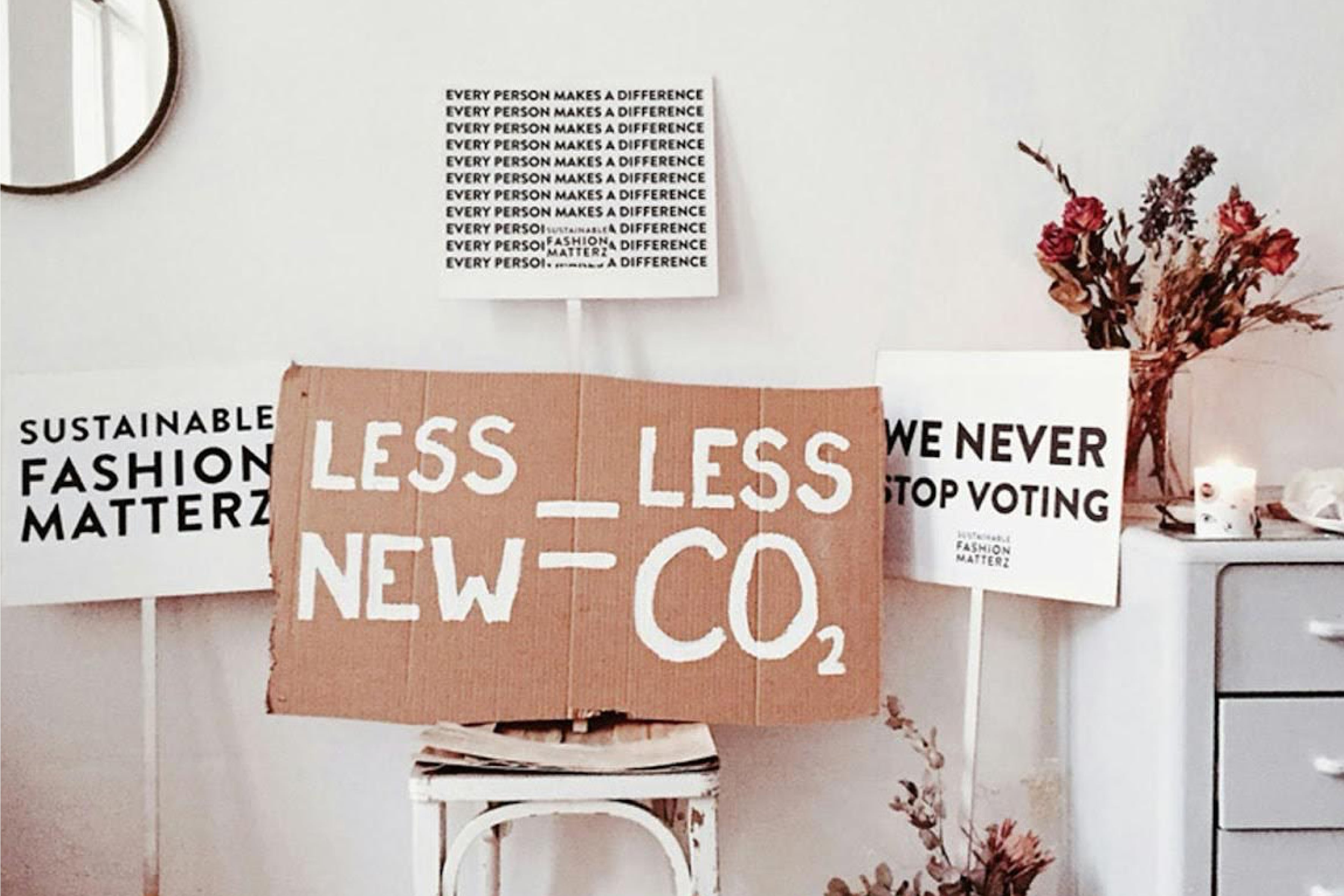 Signs with slogans about sustainability and environmental activism, including “LESS NEW = LESS CO₂” on cardboard, “Sustainable fashion matterz” and “We never stop voting,” arranged in a domestic setting with dried flowers and candles