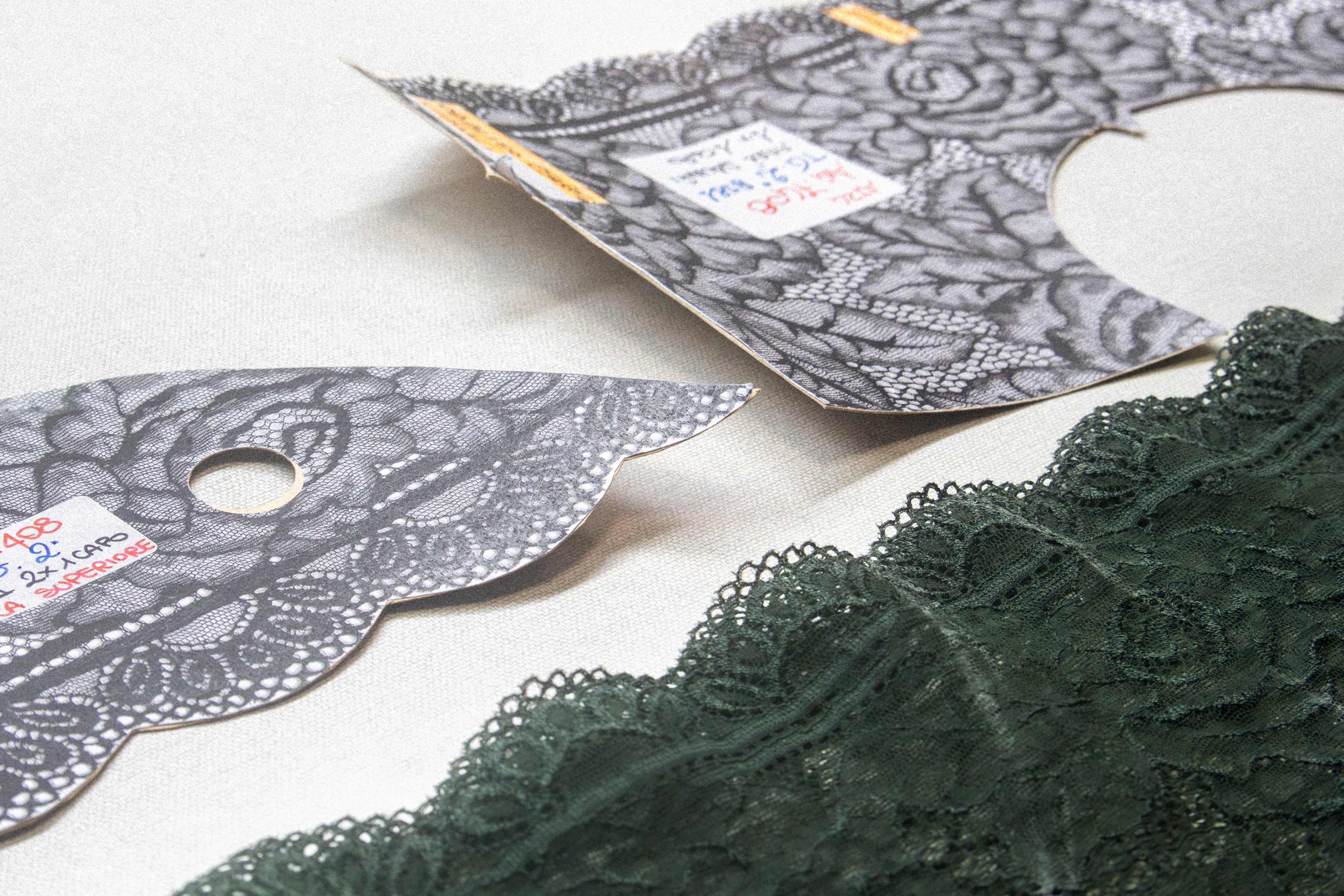 Sample of dark green Leavers lace paired with shaped cards featuring motifs and reference codes for lace production