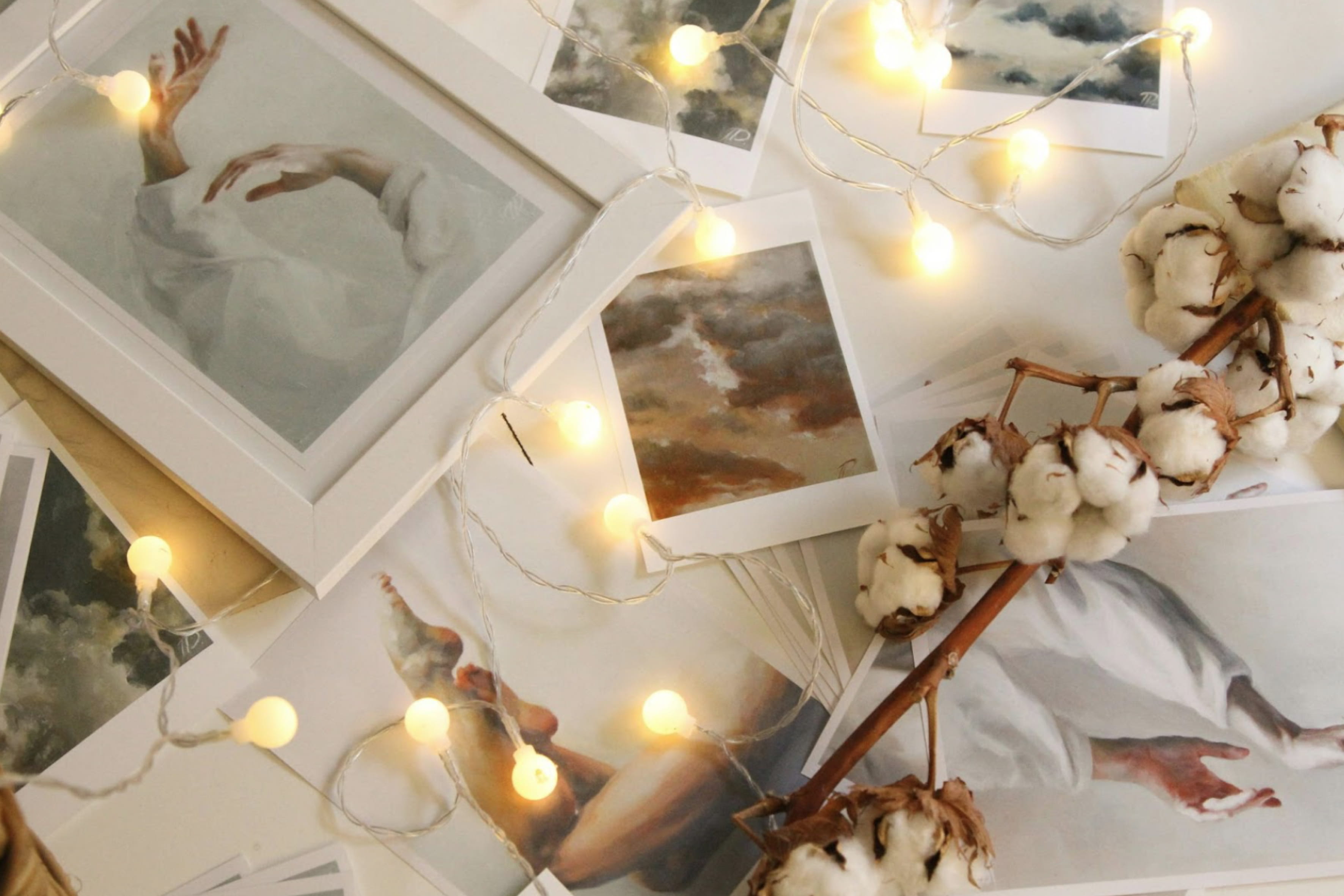 Artistic prints of hands and cloudy skies arranged on a table, decorated with warm LED lights and a cotton branch.